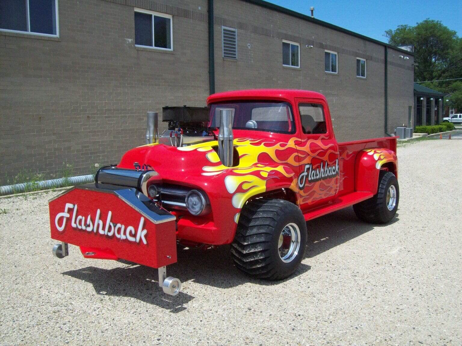 Red vintage truck with flame decals and large tires parked outside a building.