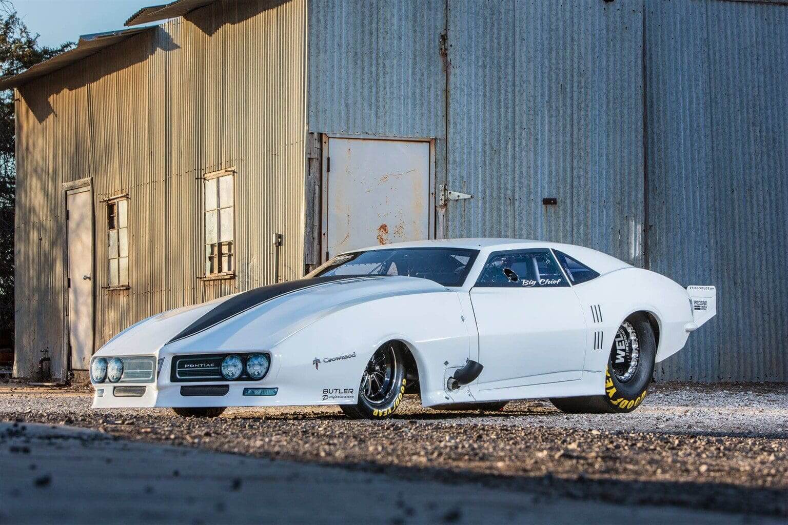 A sleek white classic muscle car parked by a rustic building.